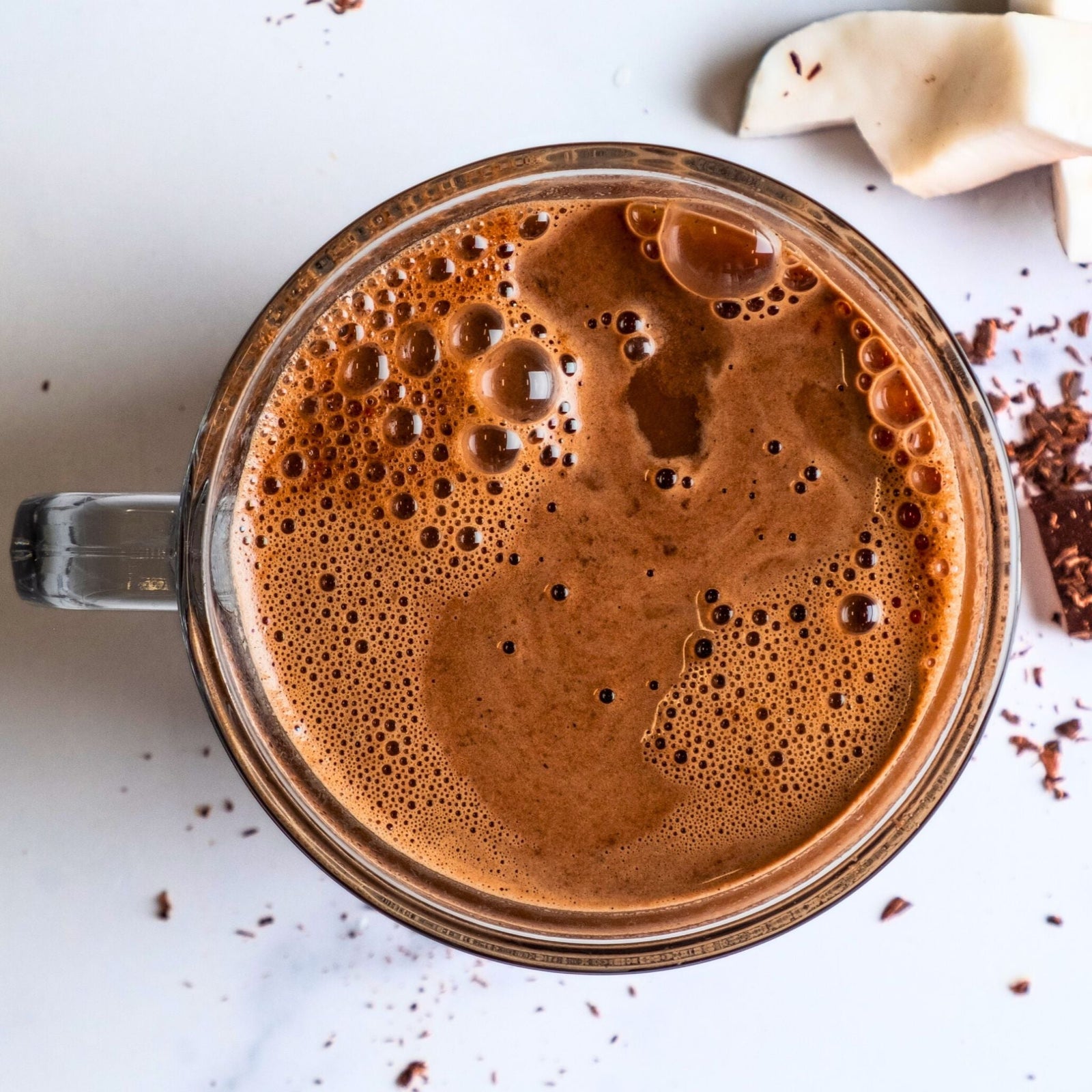 Glass mug of Bare Life's dairy-free, gluten-free, vegan hot cocoa with a frothy top, surrounded by coconut chunks and chocolate shavings on a white background.