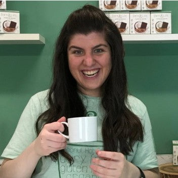  A woman enjoys a hot cocoa cup while standing before a shelf lined with assorted items.