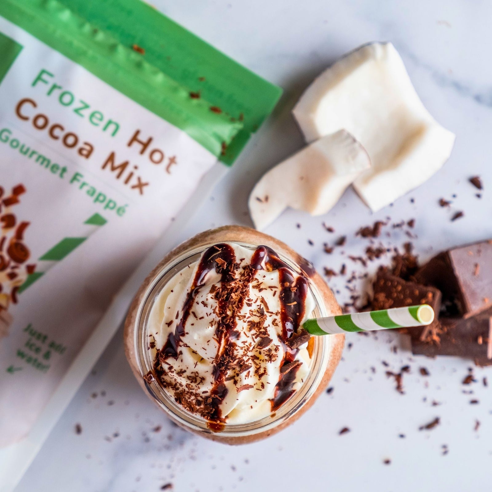 Glass of dairy-free frozen hot cocoa topped with whipped cream and chocolate drizzle, next to a package of Bare Life Frozen Hot Cocoa Mix, coconut pieces, and chocolate chunks on a white background.