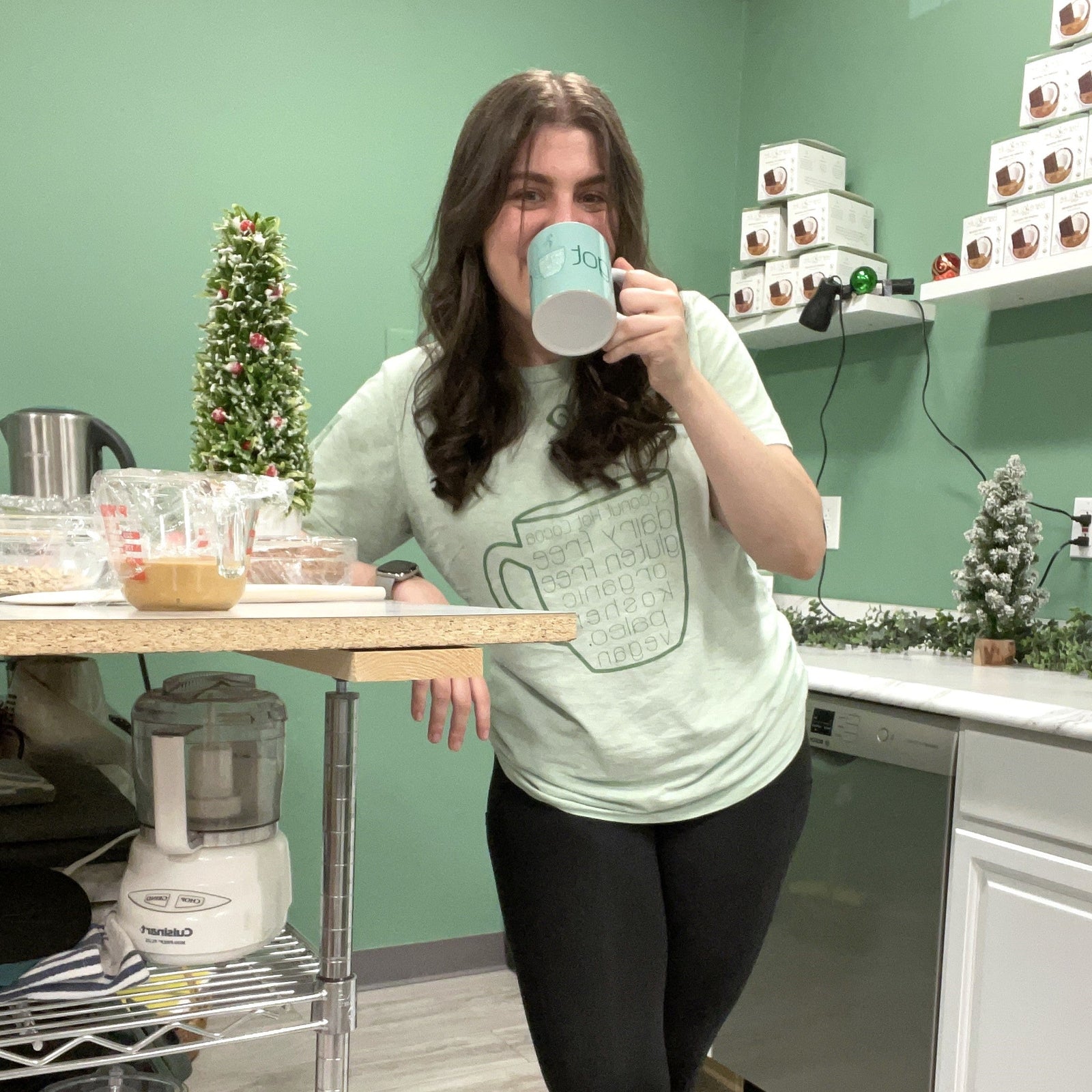 Ali Lazowski drinking from a Hot Cocoa mug in a kitchen setting with green walls and white cabinets.