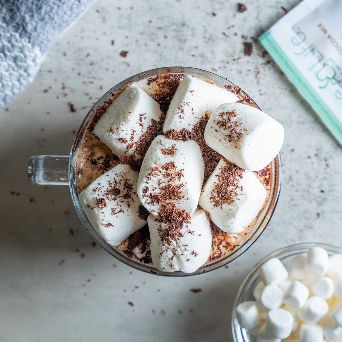Overhead view of coconut hot cocoa topped with marshmallows and chocolate shavings beside a Bare Life packet