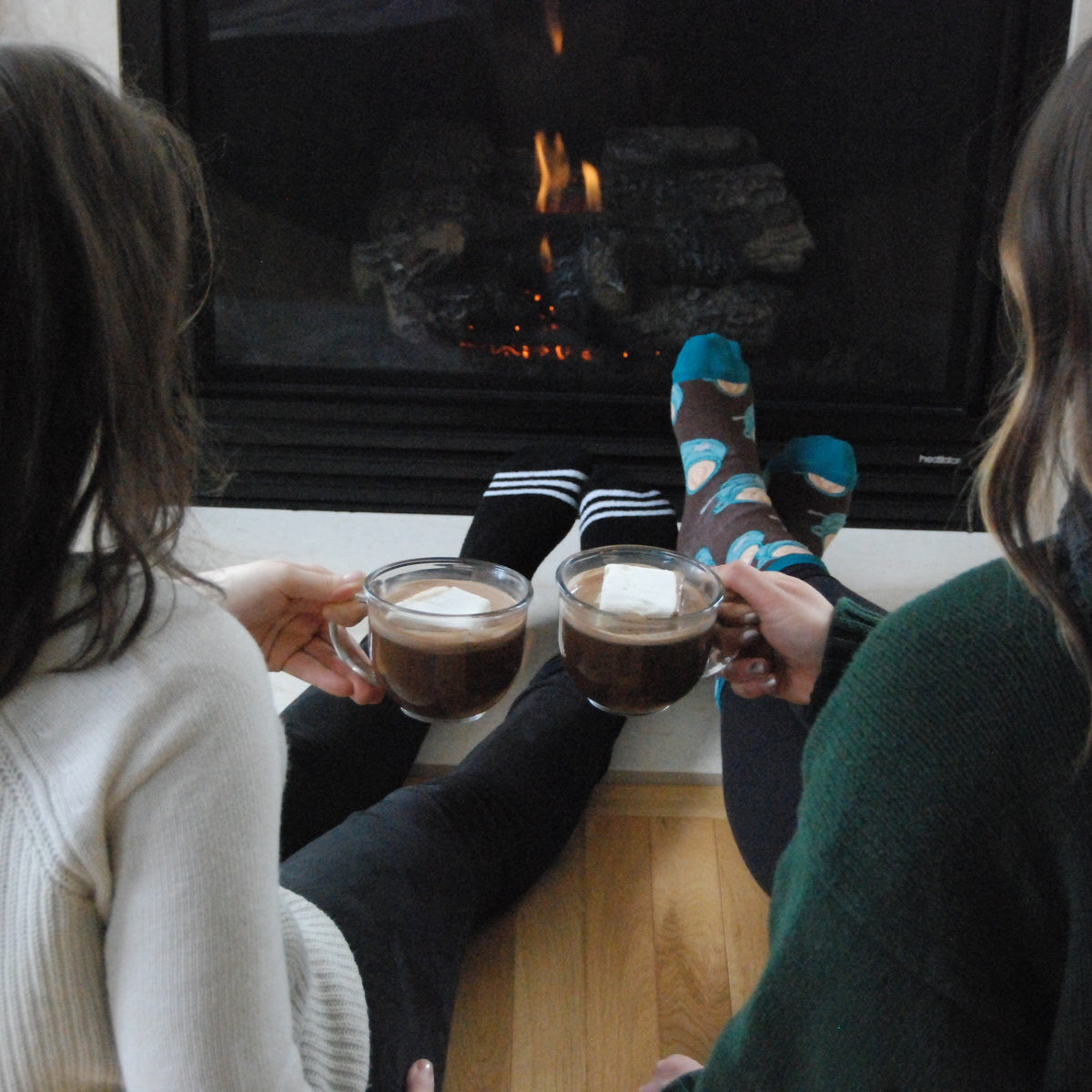 Two people holding glass mugs of hot cocoa with marshmallows while sitting beside a fireplace