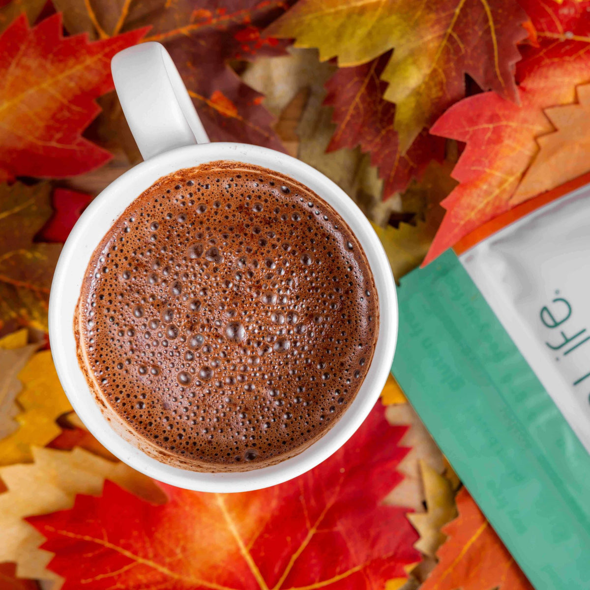 Overhead view of pumpkin spice hot cocoa in a white mug beside a Bare Life pouch and autumn leaves