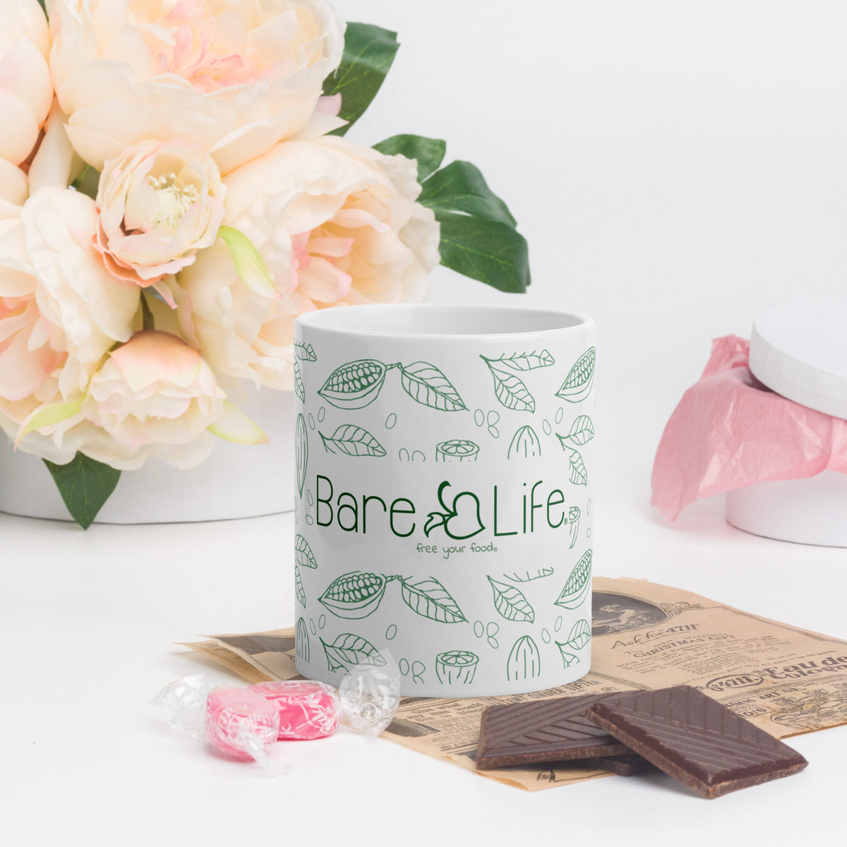 White Bare Life ceramic mug beside pale pink flowers, chocolate squares, and wrapped candy