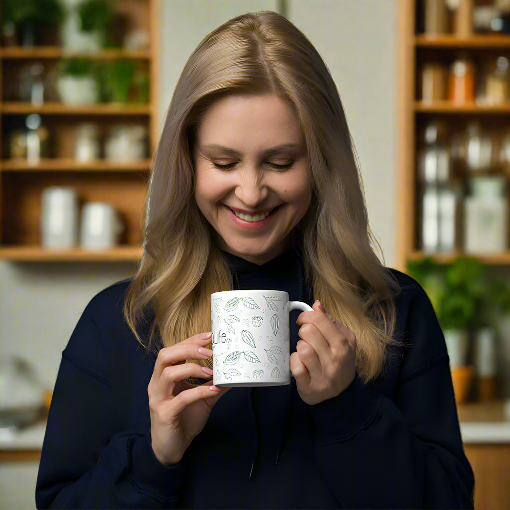 Smiling woman holding a white Bare Life ceramic mug with green cocoa pod and leaf illustrations in a kitchen